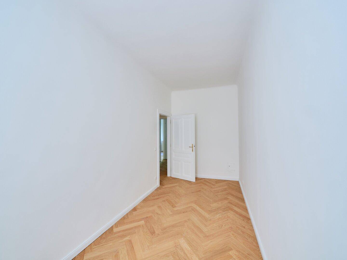 Kleiner Raum mit Fischgrätparkett und einer offenen Tür zu einem weiteren Bereich der Wohnung.