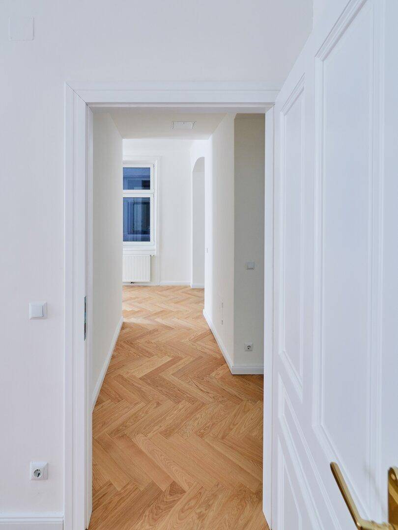 Flur mit Fischgrätparkett und Blick in einen hellen Raum mit Fenster, der viel Tageslicht bietet.