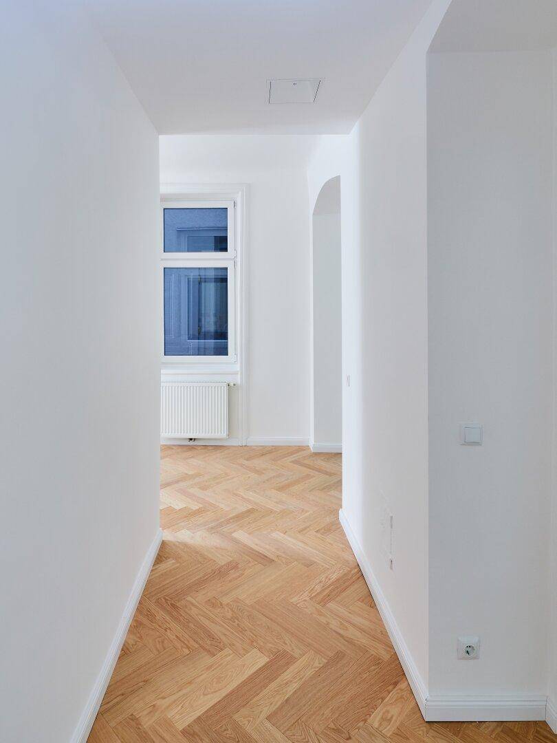 Heller Flur mit Fischgrätparkett und Blick auf ein Fenster am Ende des Ganges, das Licht spendet.