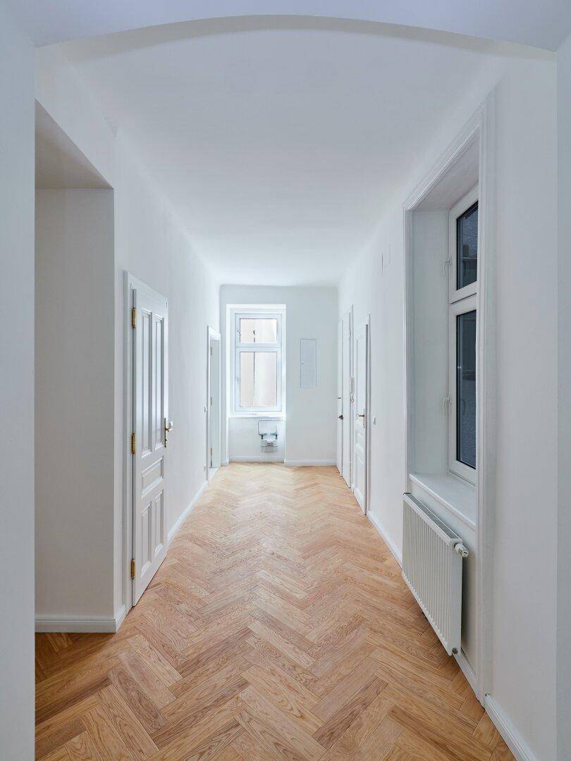Langer Flur mit Fischgrätparkett, weißen Türen und Fenstern, die den Gang mit Licht durchfluten.