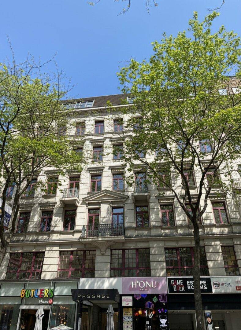 Historisches Gebäude mit Geschäften im Erdgeschoss und Wohnungen darüber, umgeben von Bäumen.