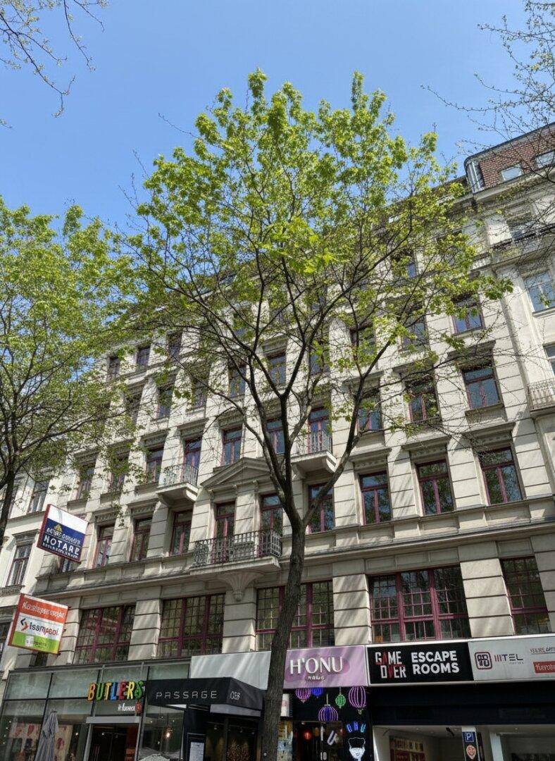 Historisches Gebäude mit Geschäften im Erdgeschoss und Wohnungen darüber, umgeben von Bäumen.