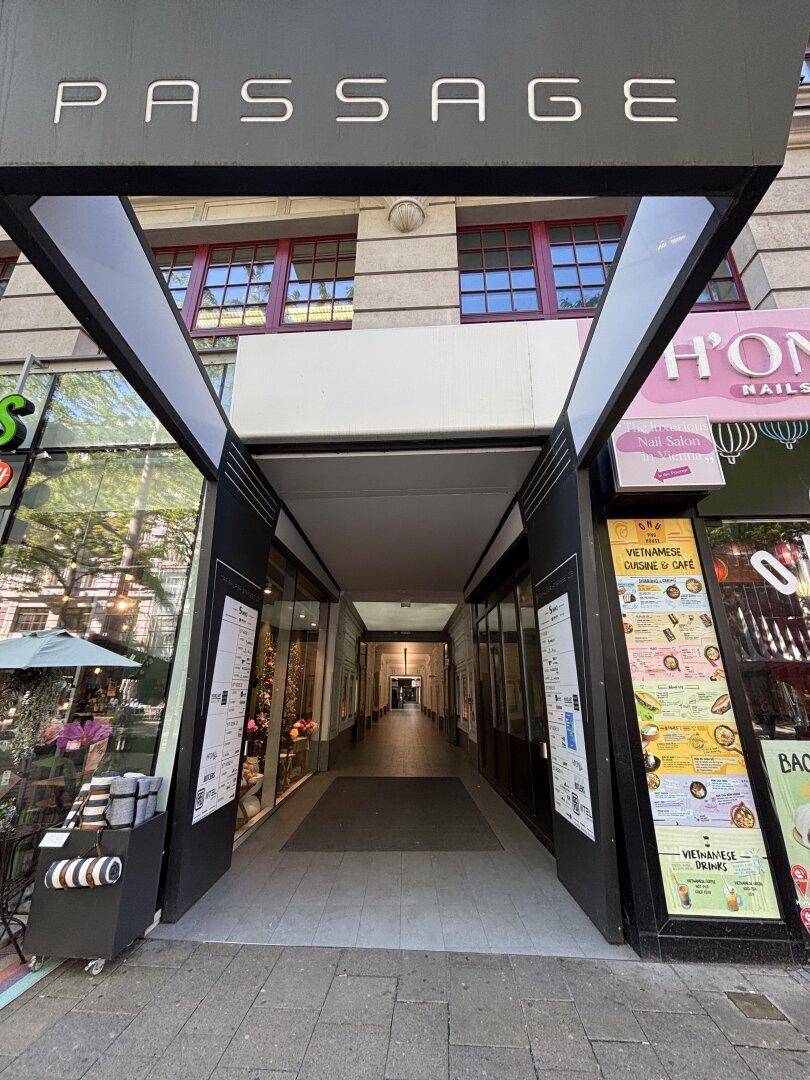 Eingang zu einer Ladenpassage mit Geschäften und Restaurants, die in einen überdachten Gang führt.