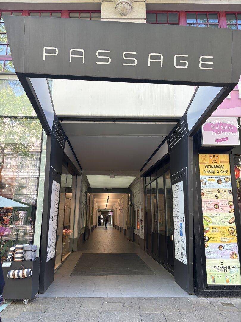 Eingang zu einer Ladenpassage mit Geschäften und Restaurants, die in einen überdachten Gang führt.