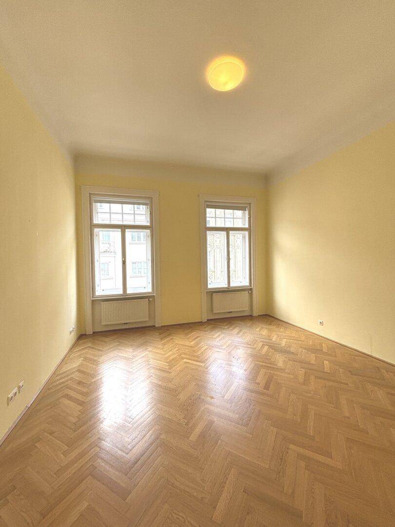 Geräumiges Zimmer mit Fischgrätparkett und zwei großen Fenstern, bietet viel Tageslicht.
