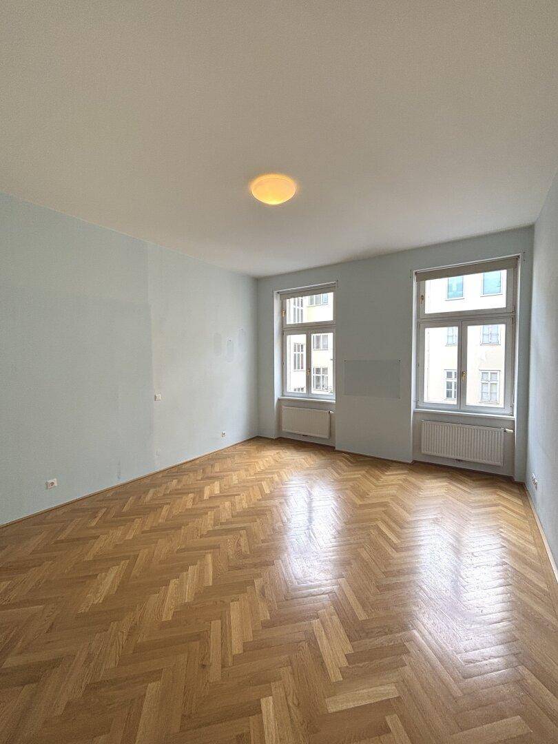 Geräumiges Zimmer mit Fischgrätparkett und zwei großen Fenstern, bereit zur individuellen Einrichtung.