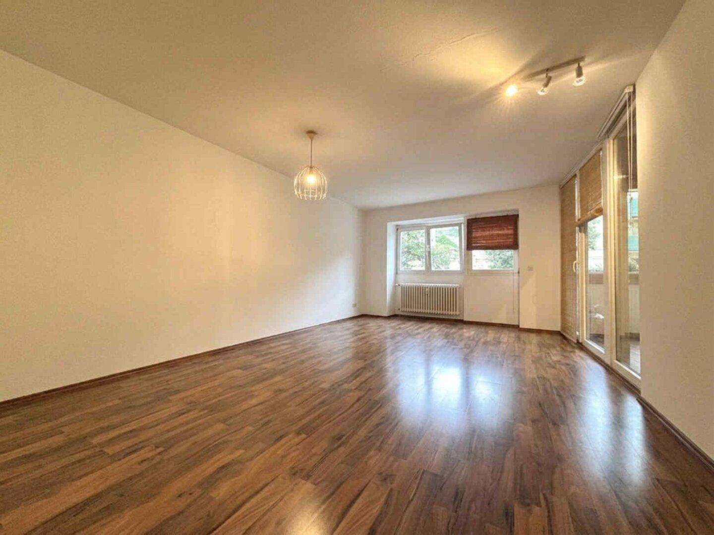 Helles Wohnzimmer mit Holzboden, Fenster und Zugang zu einem Balkon.