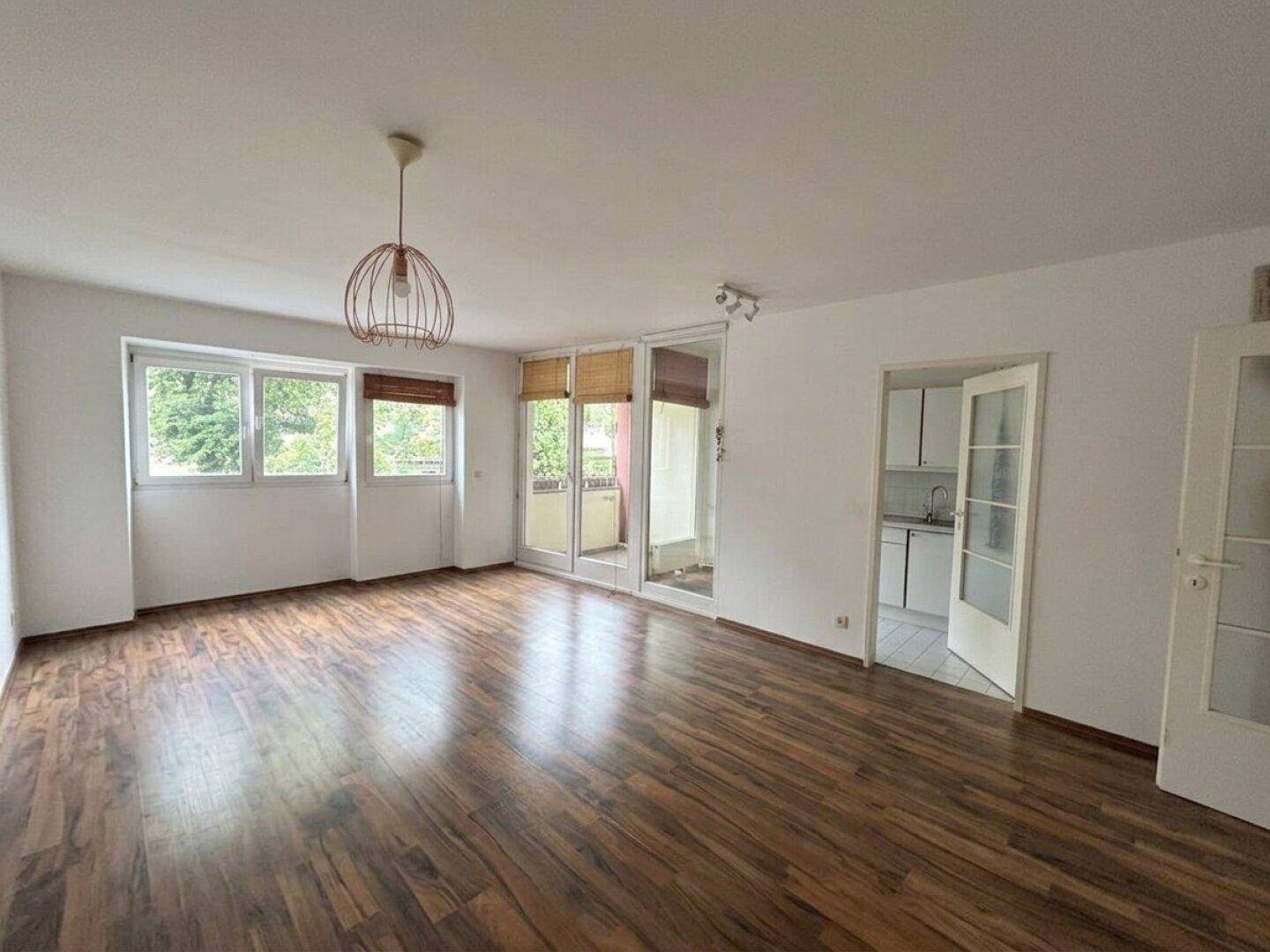 Geräumiges Wohnzimmer mit Holzboden, großen Fenstern und Zugang zum Balkon.