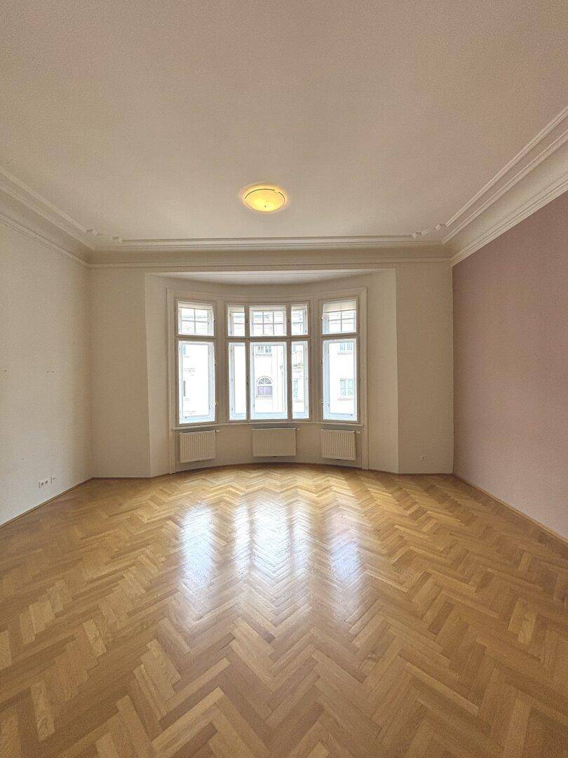 Großzügiges Zimmer mit Fischgrätparkett, Stuckdecken und einem markanten Erkerfenster.
