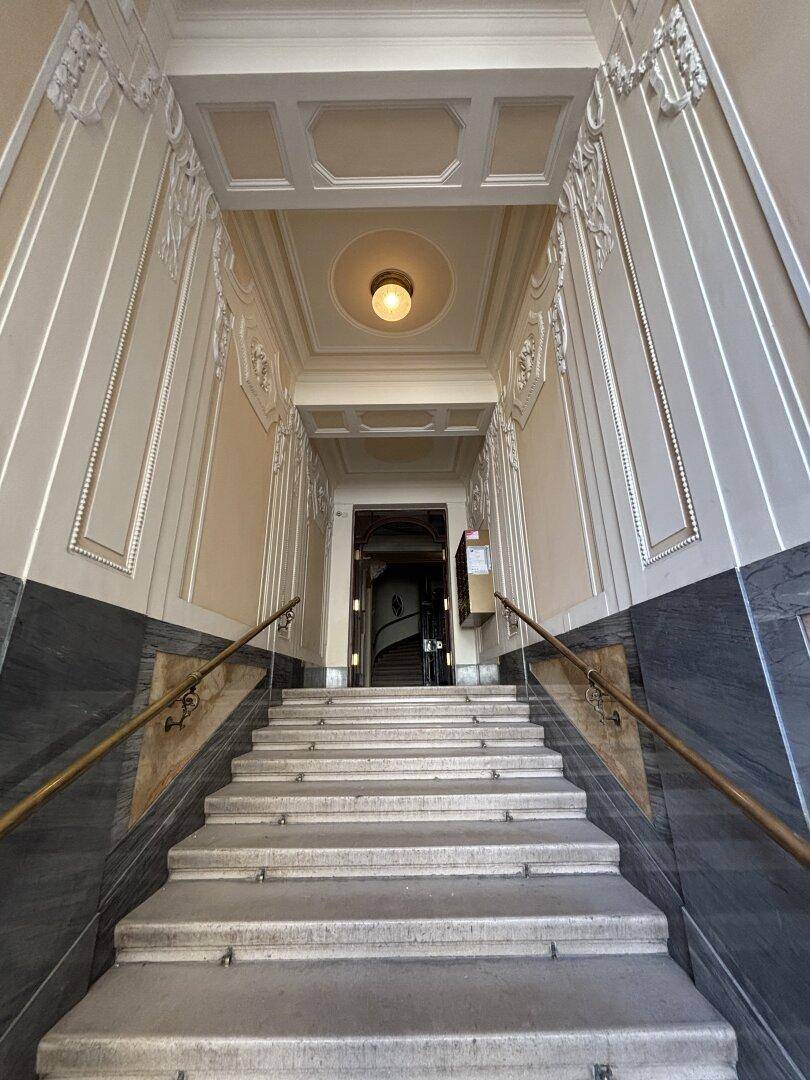 Großzügiges Treppenhaus mit Marmorstufen, hohen Decken und aufwendigen Stuckverzierungen, die zum Eingang führen.