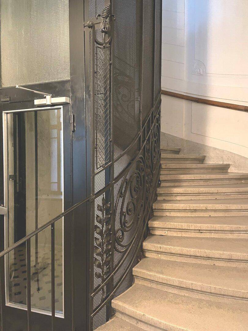 Historisches Treppenhaus mit Steinstufen und einem kunstvollen schmiedeeisernen Geländer, daneben ein Aufzug.