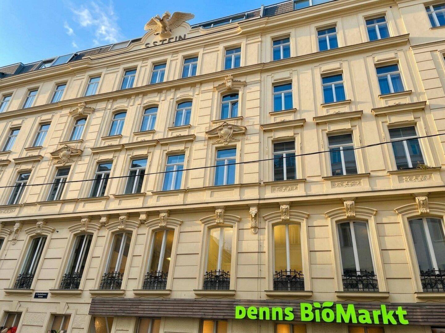 Klassisches Altbaugebäude mit vielen Fenstern und einem Geschäft im Erdgeschoss, unter blauem Himmel.