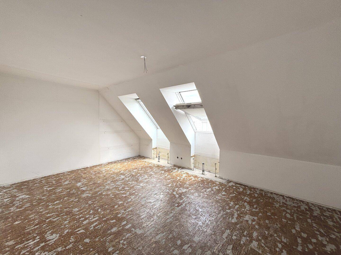 Dachgeschossraum mit schrägen Wänden, Dachfenstern und sichtbaren Installationen, bereit für den Ausbau.