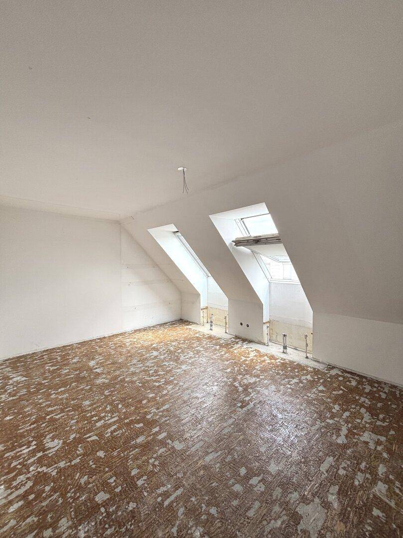 Geräumiger Dachgeschossraum mit schrägen Wänden und Dachfenstern, der auf die Fertigstellung wartet.