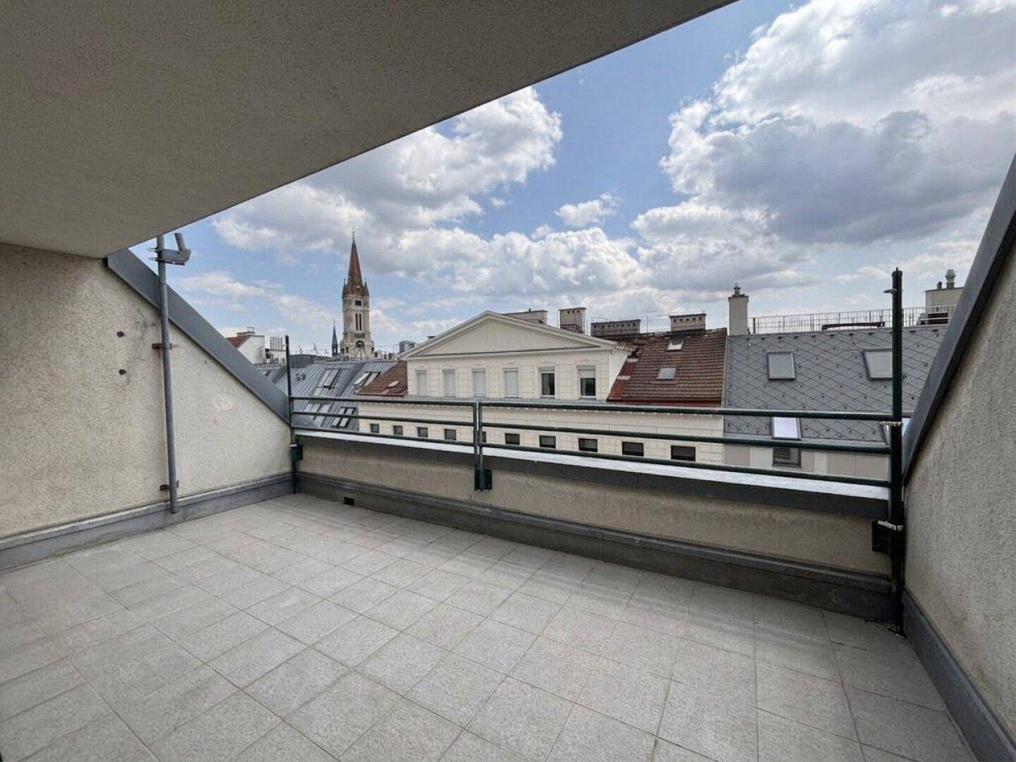 Geräumiger Balkon mit Fliesenboden und weitem Blick über die Dächer der Stadt und einen Kirchturm.