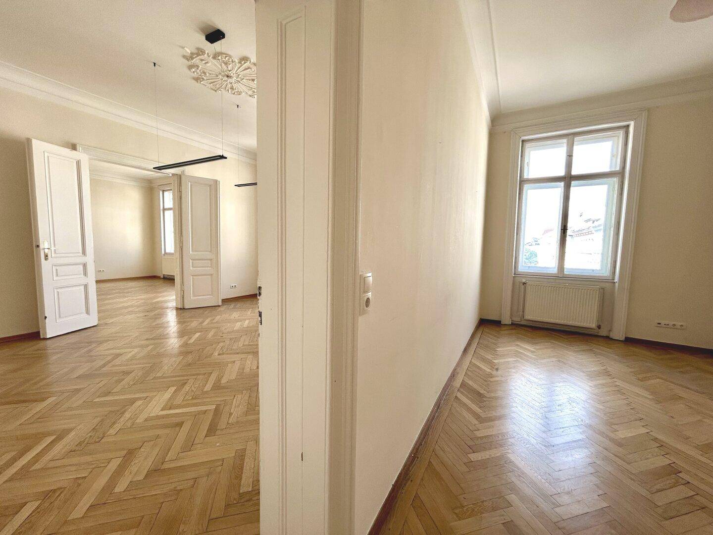 Helles Wohnzimmer mit Fischgrätparkett, hohen Decken und Stuckverzierung, durch große Fenster gut belichtet.