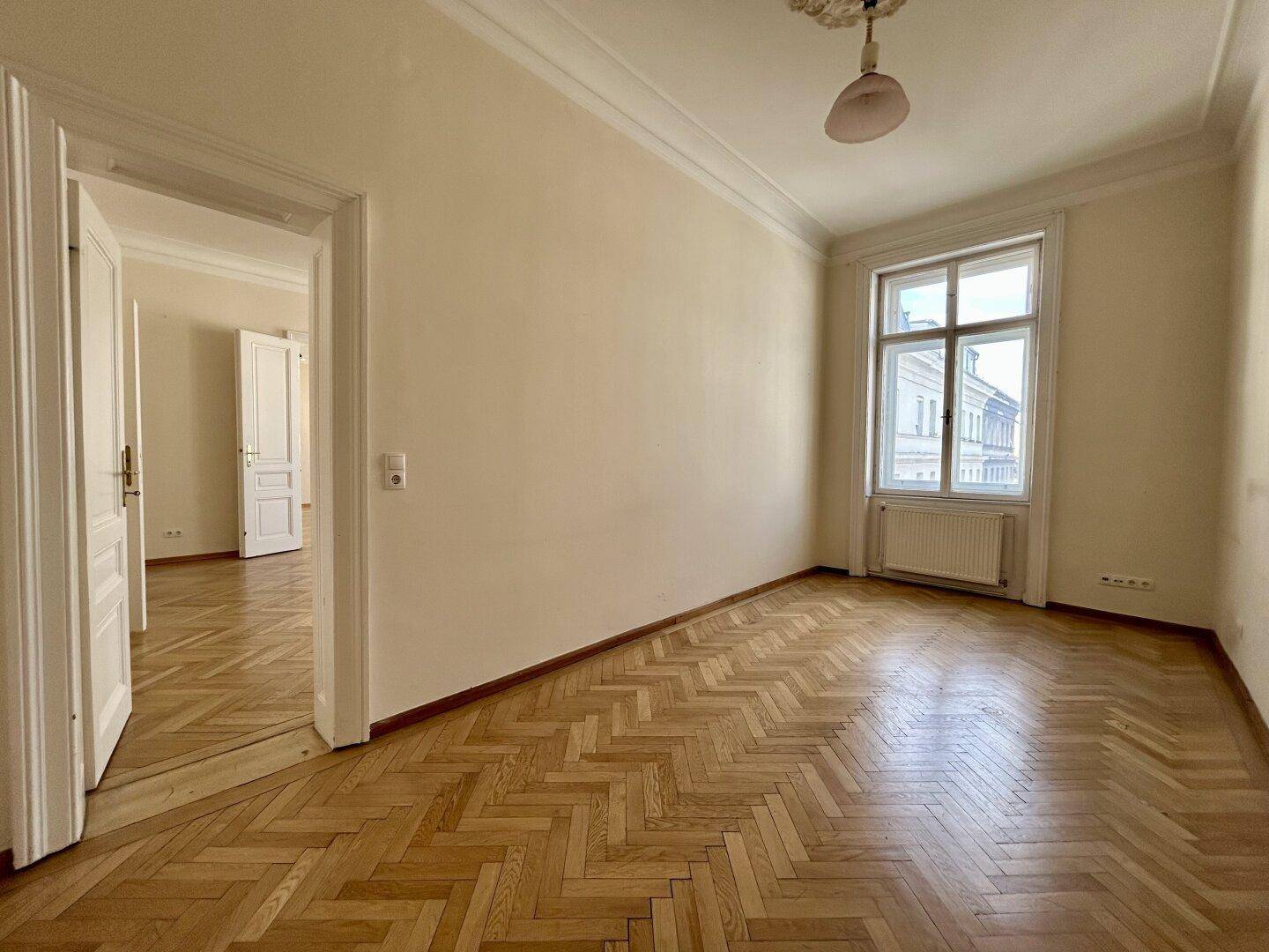 Geräumiges Zimmer mit Fischgrätparkett, hohen Decken und einem großen Fenster, das viel Licht spendet.