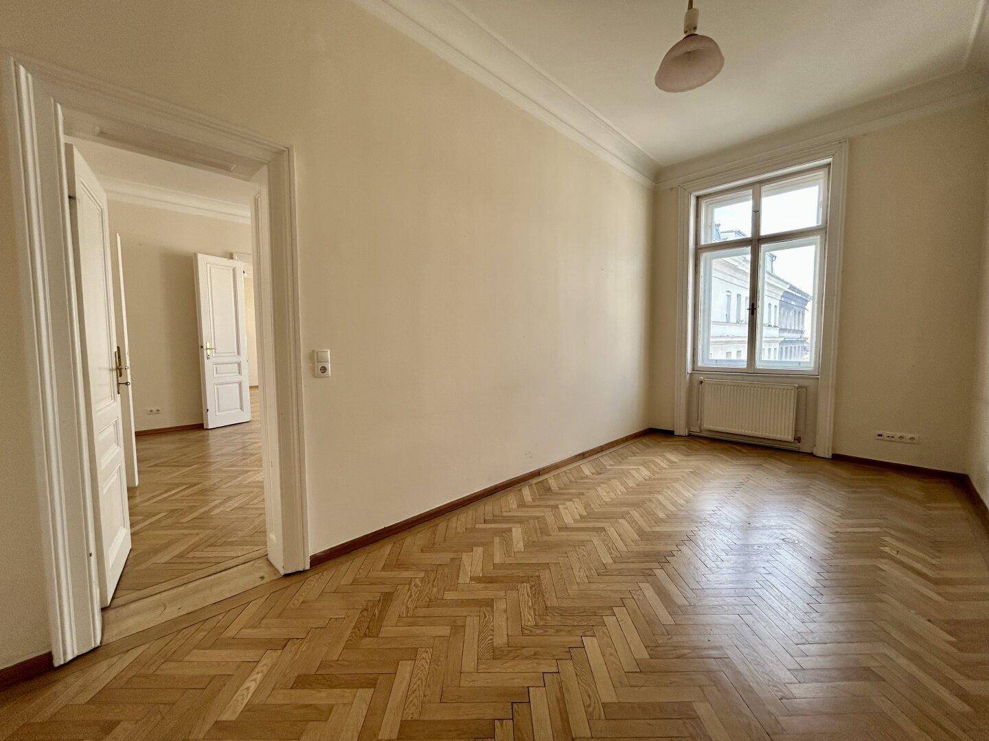 Großes Zimmer mit Fischgrätparkett, hohen Decken und einem Fenster, das den Raum mit Tageslicht füllt.