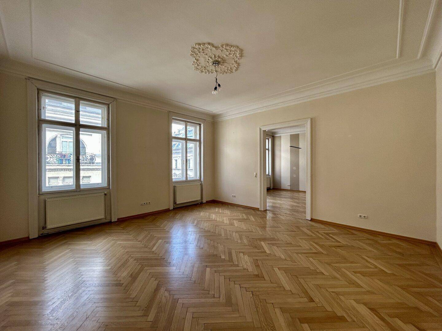 Helles Wohnzimmer mit Fischgrätparkett, hohen Decken und Stuckverzierung, durch große Fenster gut belichtet.