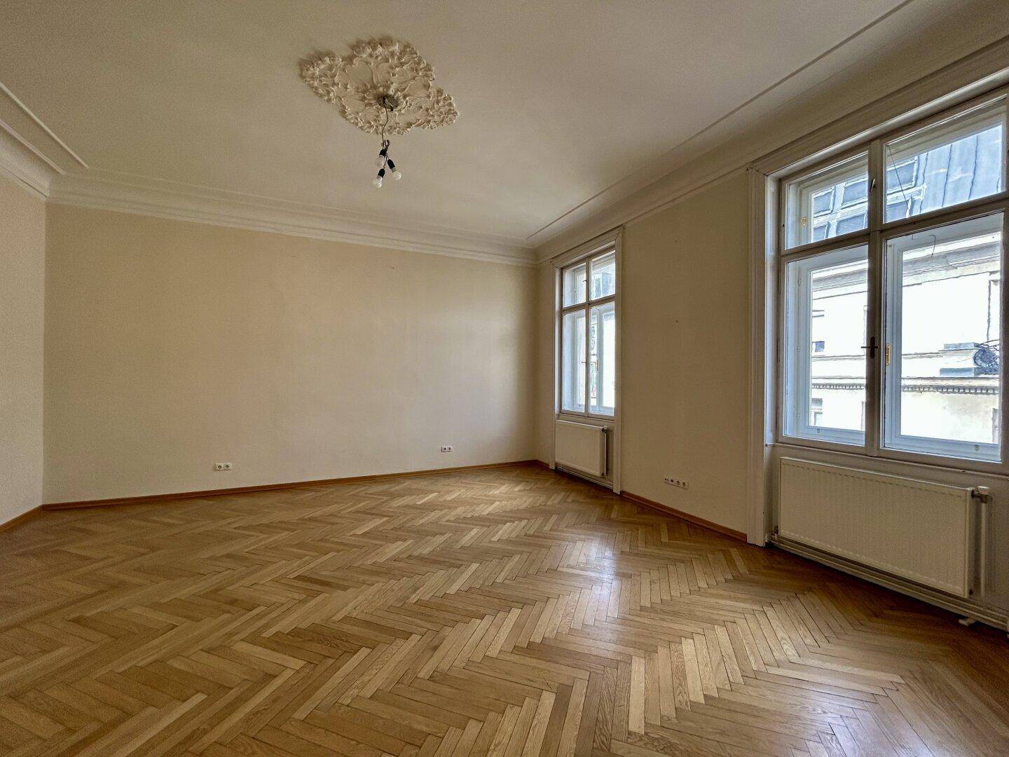 Helles Wohnzimmer mit klassischem Fischgrätparkett, hohen Decken und Stuckelementen, durch große Fenster gut belichtet.