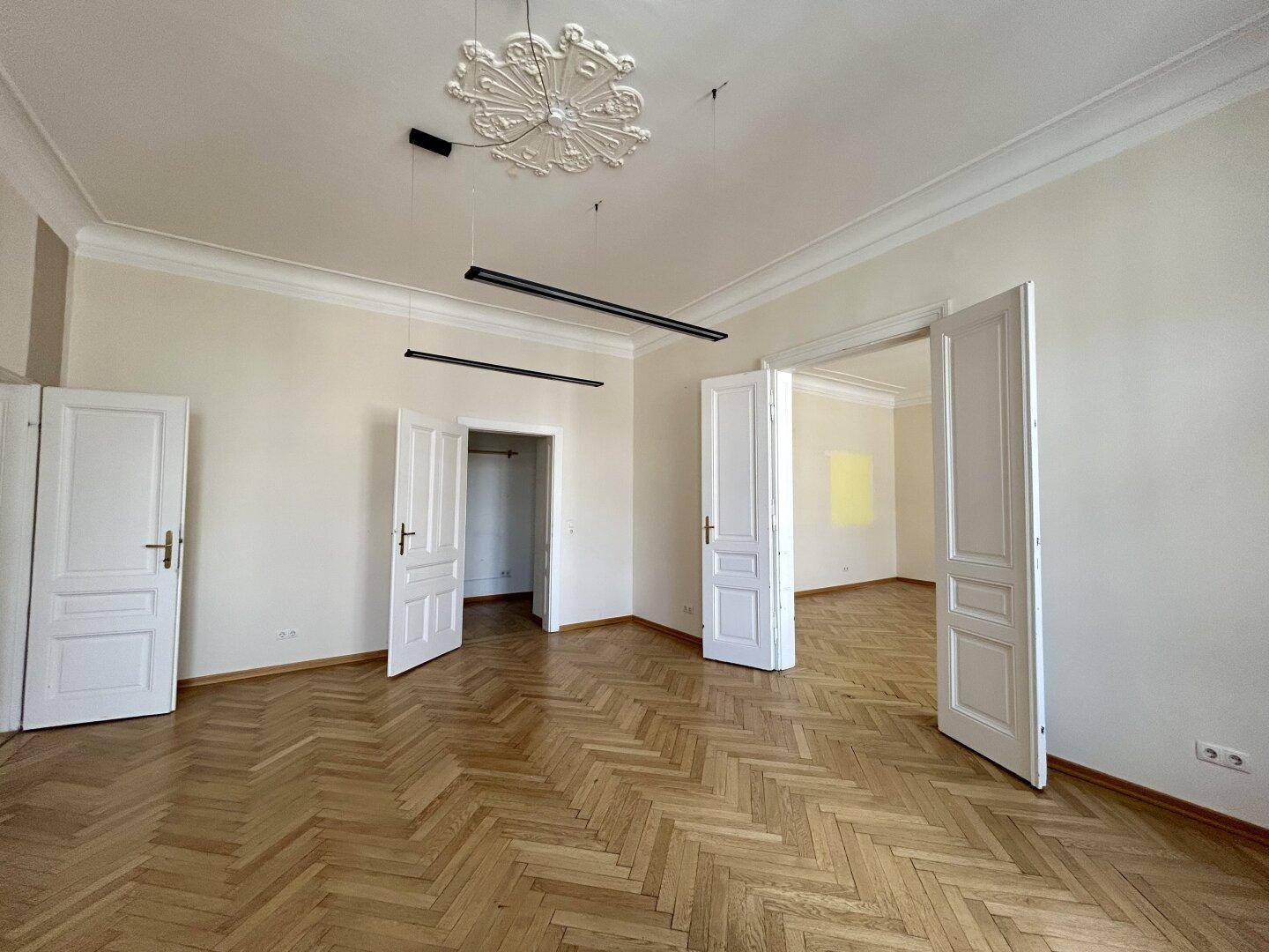 Großzügiges Wohnzimmer mit Fischgrätparkett, hohen Decken und mehreren Türen zu angrenzenden Räumen.