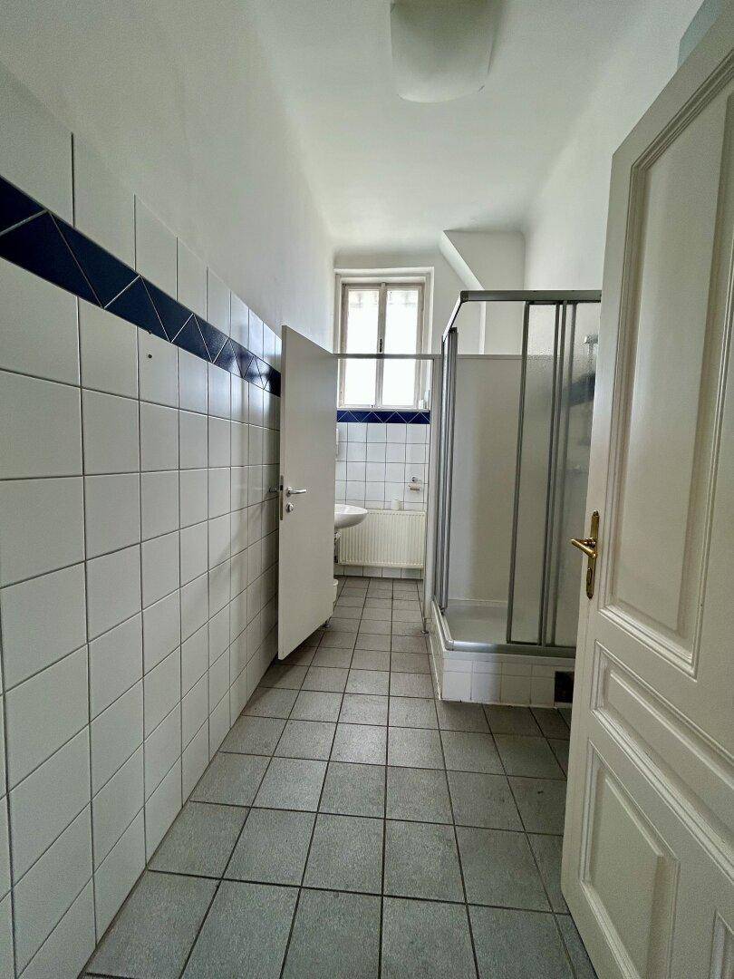 Badezimmer mit weißen Fliesen, Duschkabine und einem Fenster, das für Belüftung sorgt.