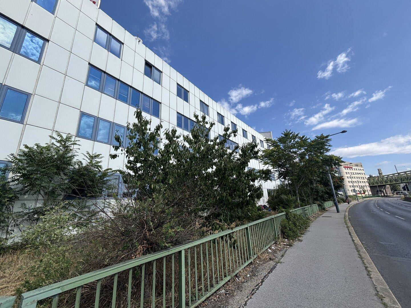 Moderne Gebäudefassade mit vielen Fenstern, umgeben von Bäumen und einer Straße.