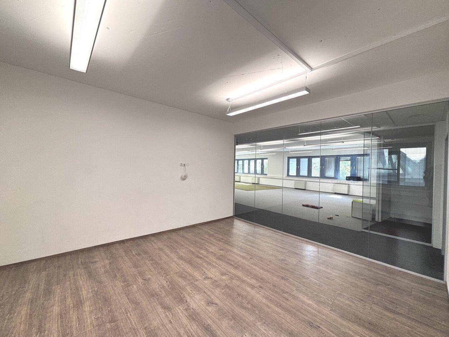 Heller Büroraum mit Laminatboden und Glaswand, die den Blick auf weitere Bürobereiche freigibt.