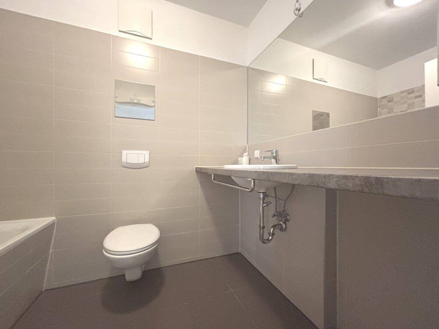 Modernes Badezimmer mit großen Fliesen, Badewanne, WC und breitem Waschtisch.
