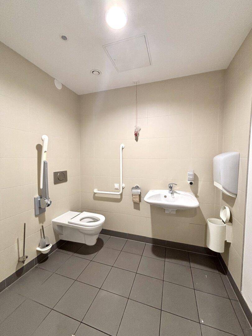 Barrierefreies Badezimmer mit Wand-WC, Haltegriffen und Waschbecken, alles in hellen Farbtönen gehalten.