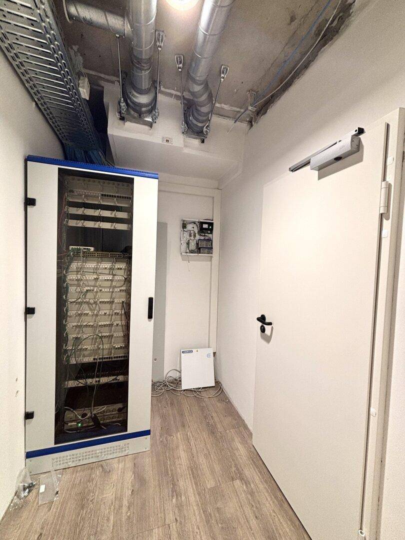 Serverraum mit einem Serverschrank, sichtbaren Lüftungsrohren und Kabeltrassen an der Decke.