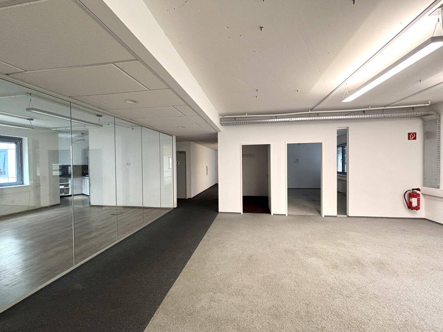 Offener Bürobereich mit Teppichboden, Glaswänden und einem langen Flur, der zu weiteren Räumen führt.