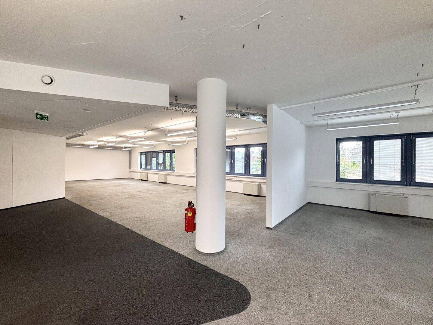 Offener Bürobereich mit Teppichboden, einer zentralen Säule und großen Fenstern mit Blick nach draußen.