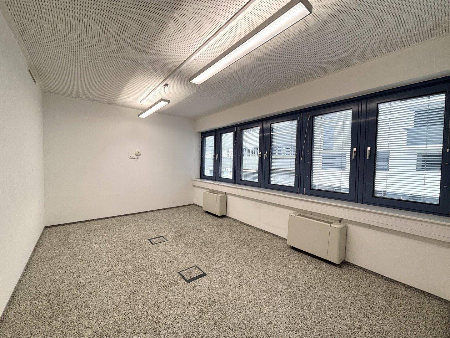 Leerer Büroraum mit Teppichboden, großen Fenstern mit Jalousien und Bodentanks für flexible Arbeitsplatzgestaltung.