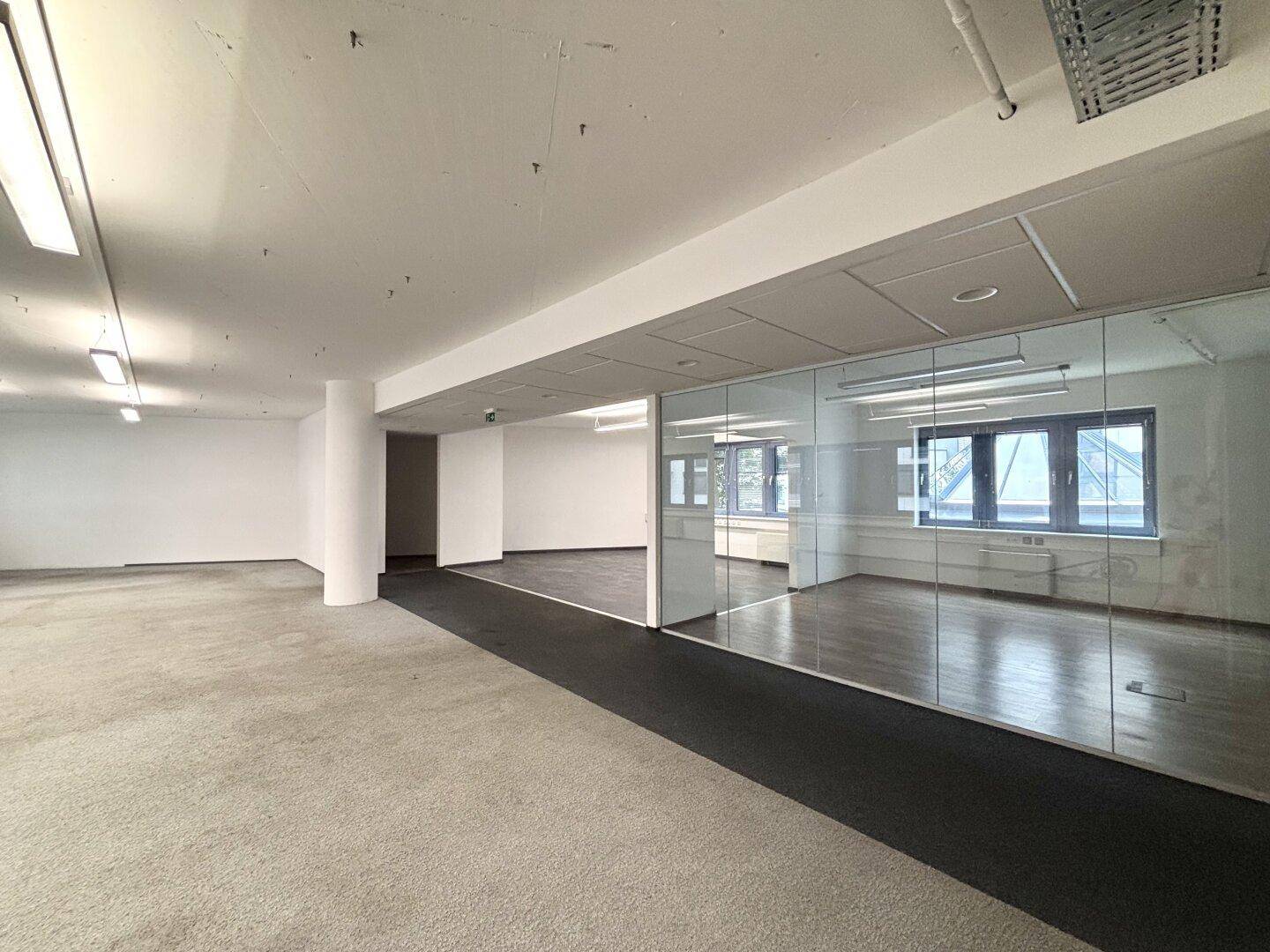 Großzügige offene Bürofläche mit Teppich- und Holzoptikboden sowie einer modernen Glasabtrennung.