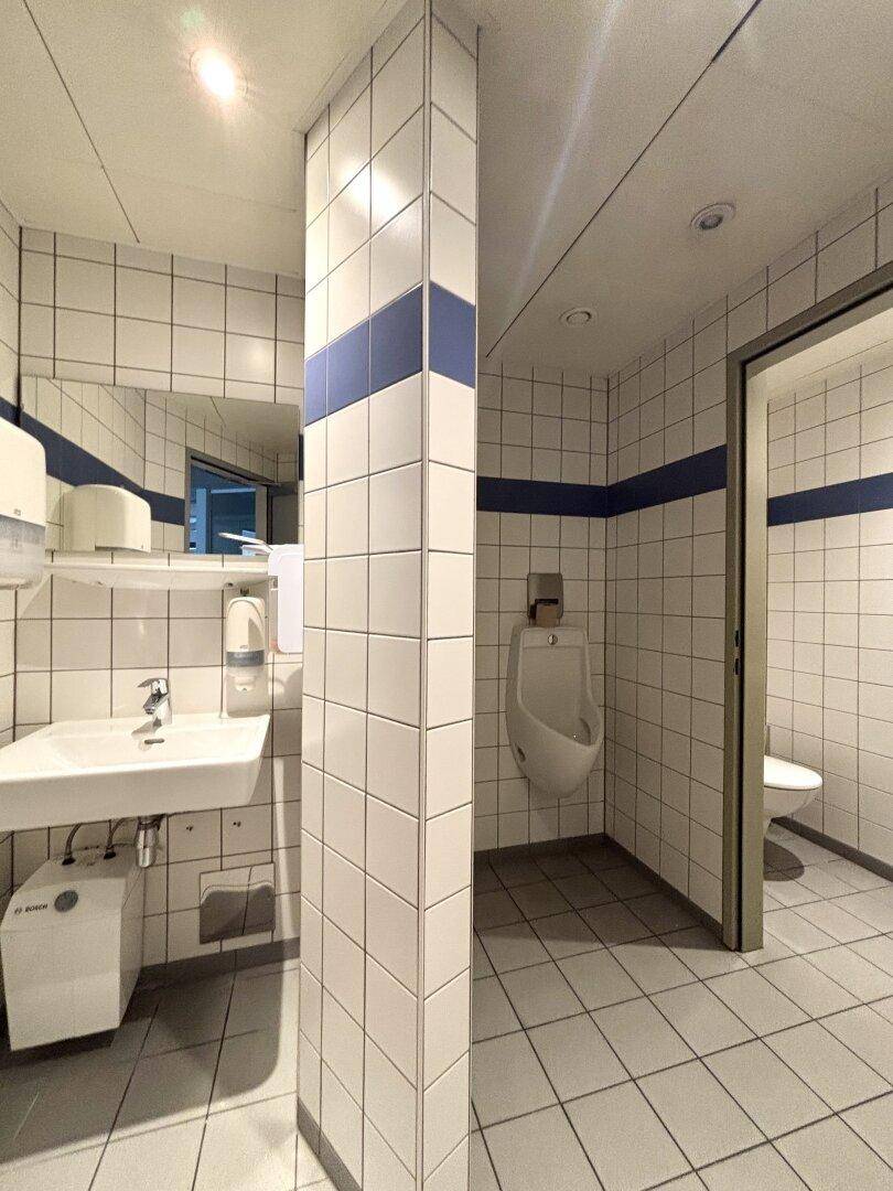 Öffentliche Toilette mit weißen Fliesen, einem Urinal, Waschbecken und einer Toilettenkabine.