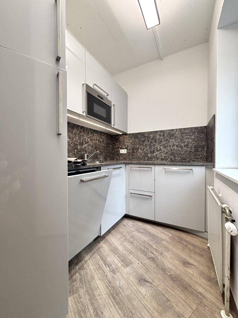 Kleine, moderne Küchenzeile mit weißen Schränken, integrierter Mikrowelle und Holzoptikboden.