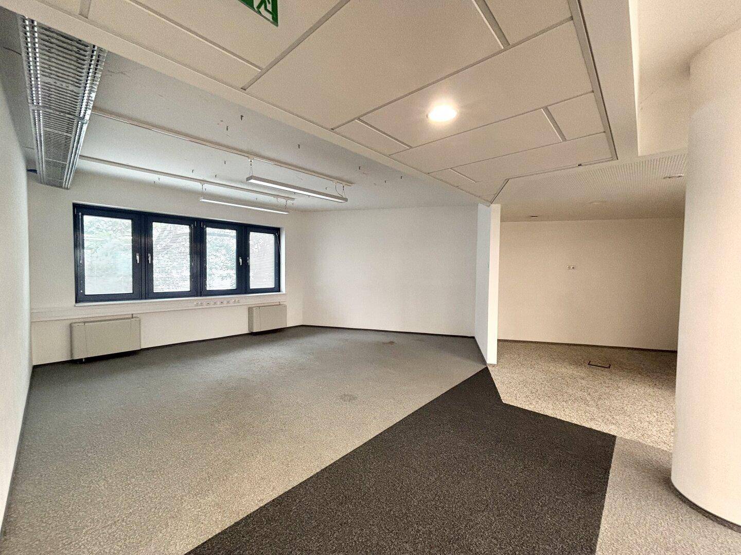 Heller Büroraum mit Teppichboden, großen Fenstern und sichtbaren Lüftungskanälen an der Decke.