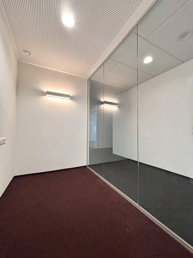 Kleiner Büroraum mit rotem Teppichboden, Glastrennwänden und moderner Wandbeleuchtung.