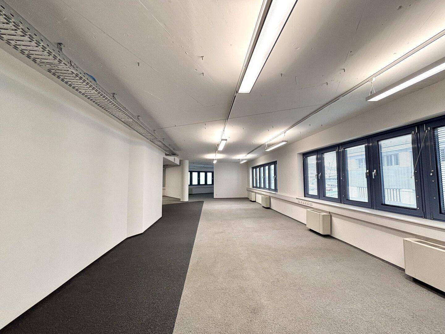 Langer, offener Bürobereich mit Teppichboden, vielen Fenstern und sichtbaren Kabelkanälen an der Decke.
