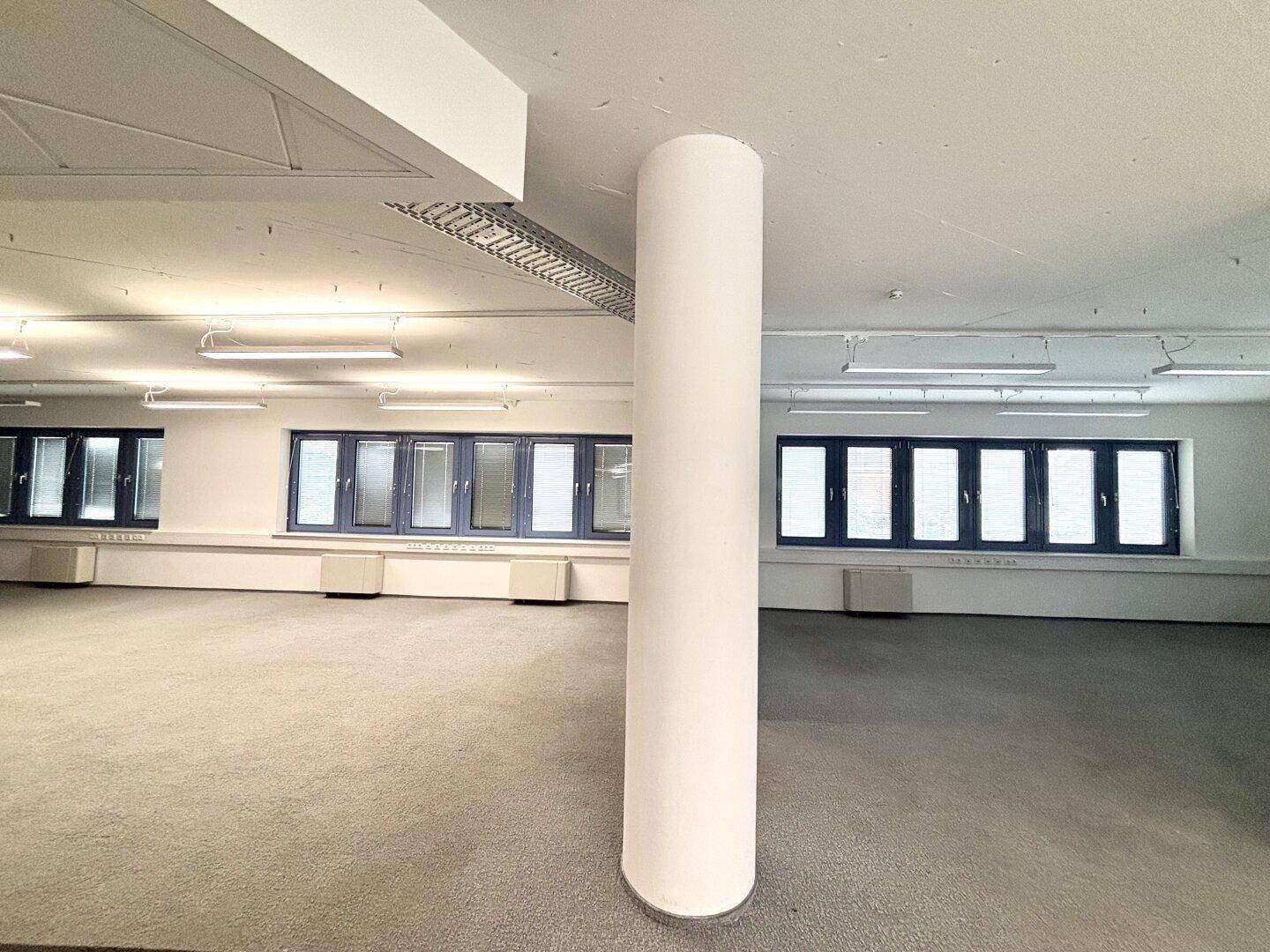 Großer Bürobereich mit zentraler Säule, Teppichboden und vielen Fenstern, die Tageslicht hereinlassen.