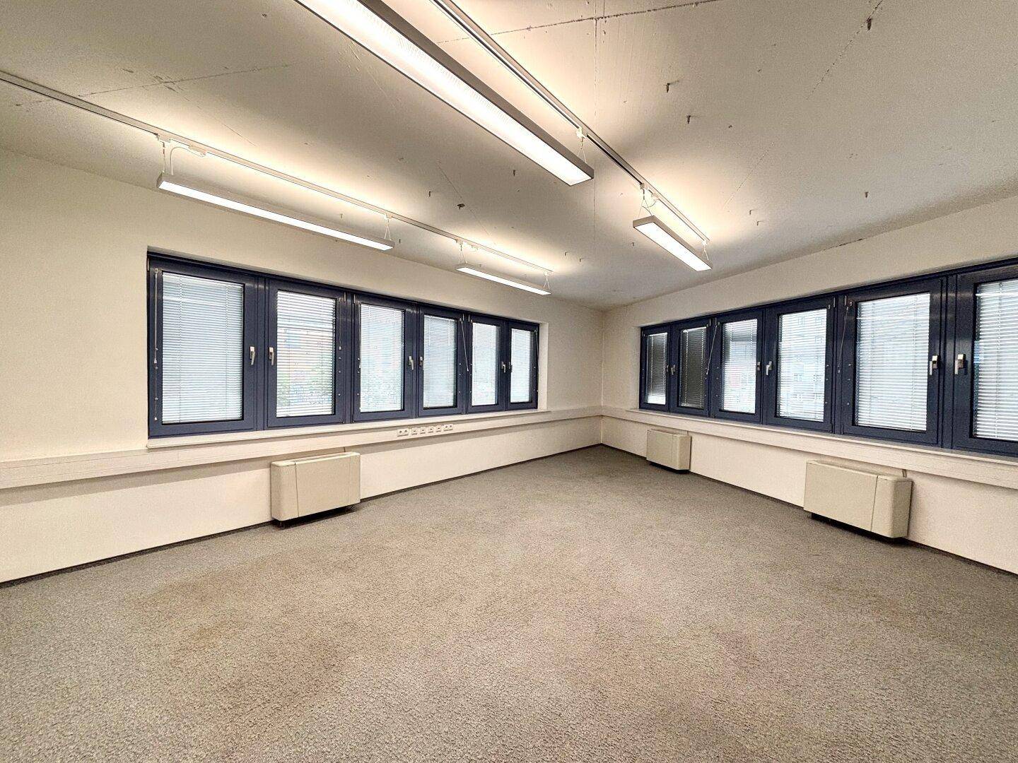 Heller Büroraum mit Teppichboden, blauen Fensterrahmen und Heizkörpern unter den Fenstern.