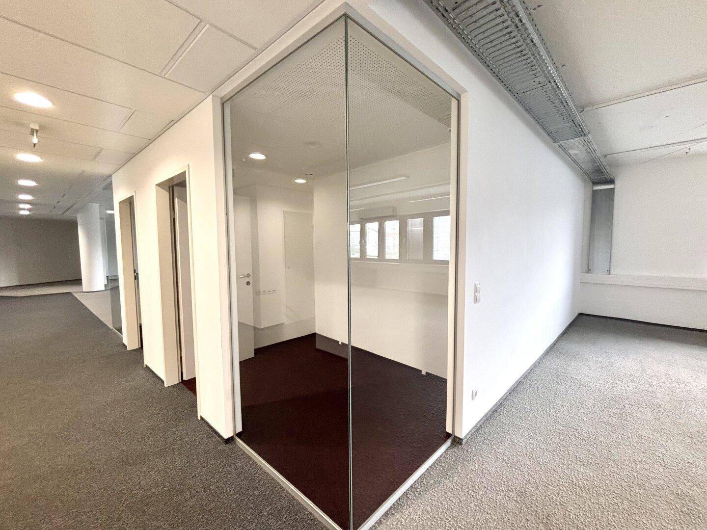 Moderner Bürobereich mit Glaswänden, die einen separaten Raum abgrenzen, und Teppichboden im Flur.