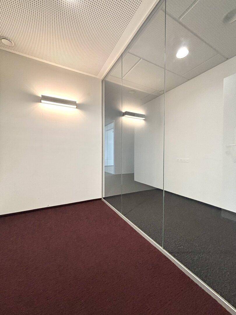 Innenansicht eines Büros mit Glaswand, rotem Teppichboden und Wandleuchten, die den Raum erhellen.