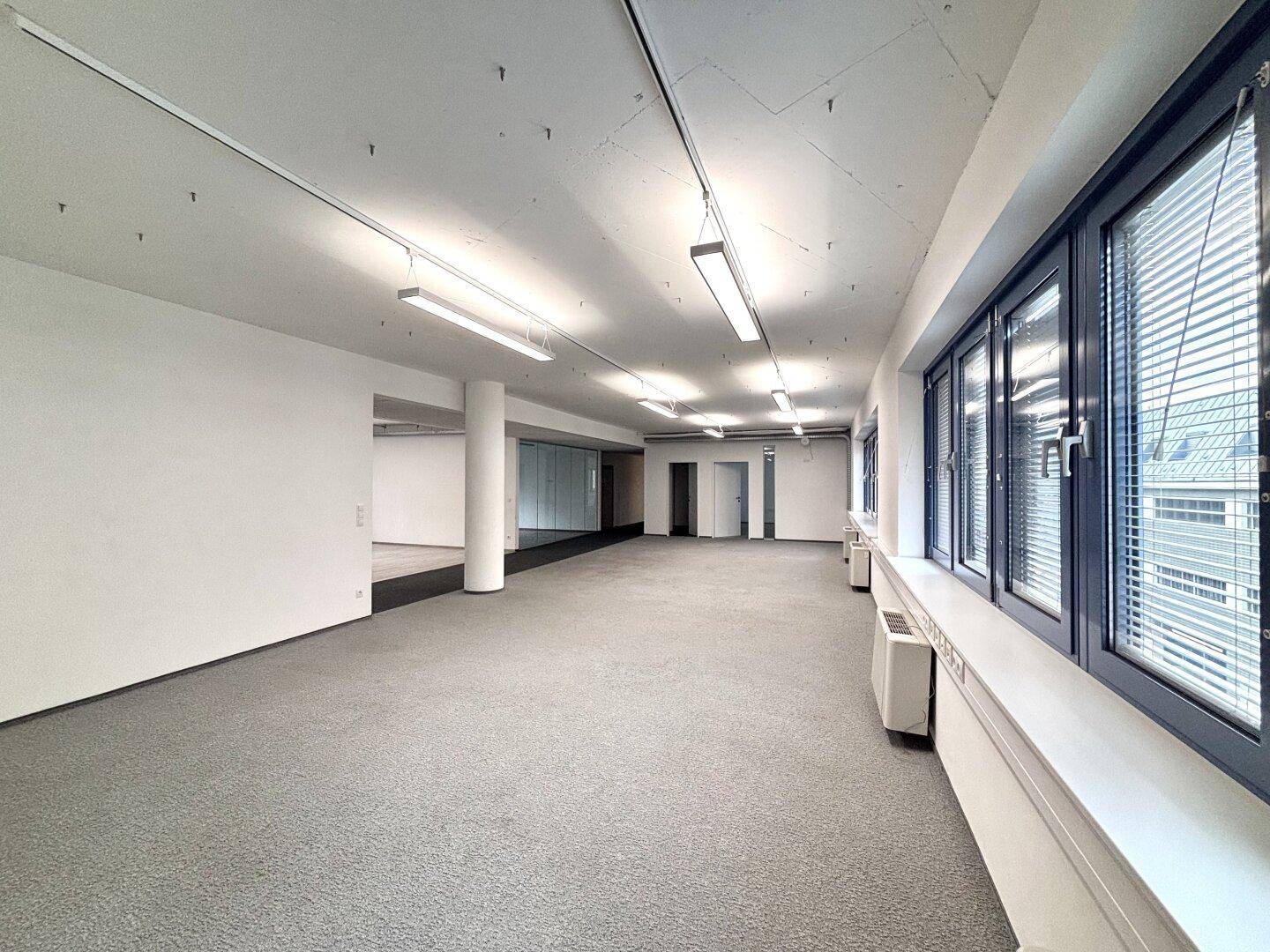 Langer Büroraum mit Teppichboden, vielen Fenstern und Deckenbeleuchtung, bereit zur individuellen Gestaltung.