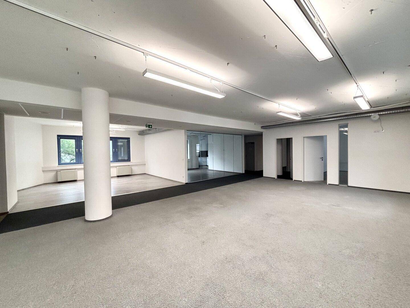 Weitläufiger Bürobereich mit Teppichboden, Säule und Glaswänden, die separate Büros abtrennen.