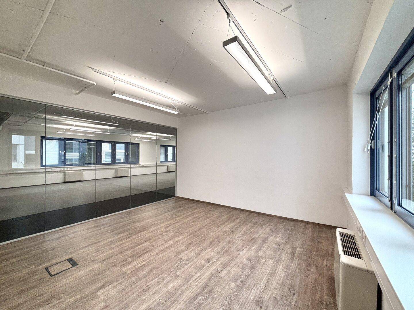 Büroraum mit Holzoptik-Boden, Glaswand und Fenstern, die viel natürliches Licht hereinlassen.