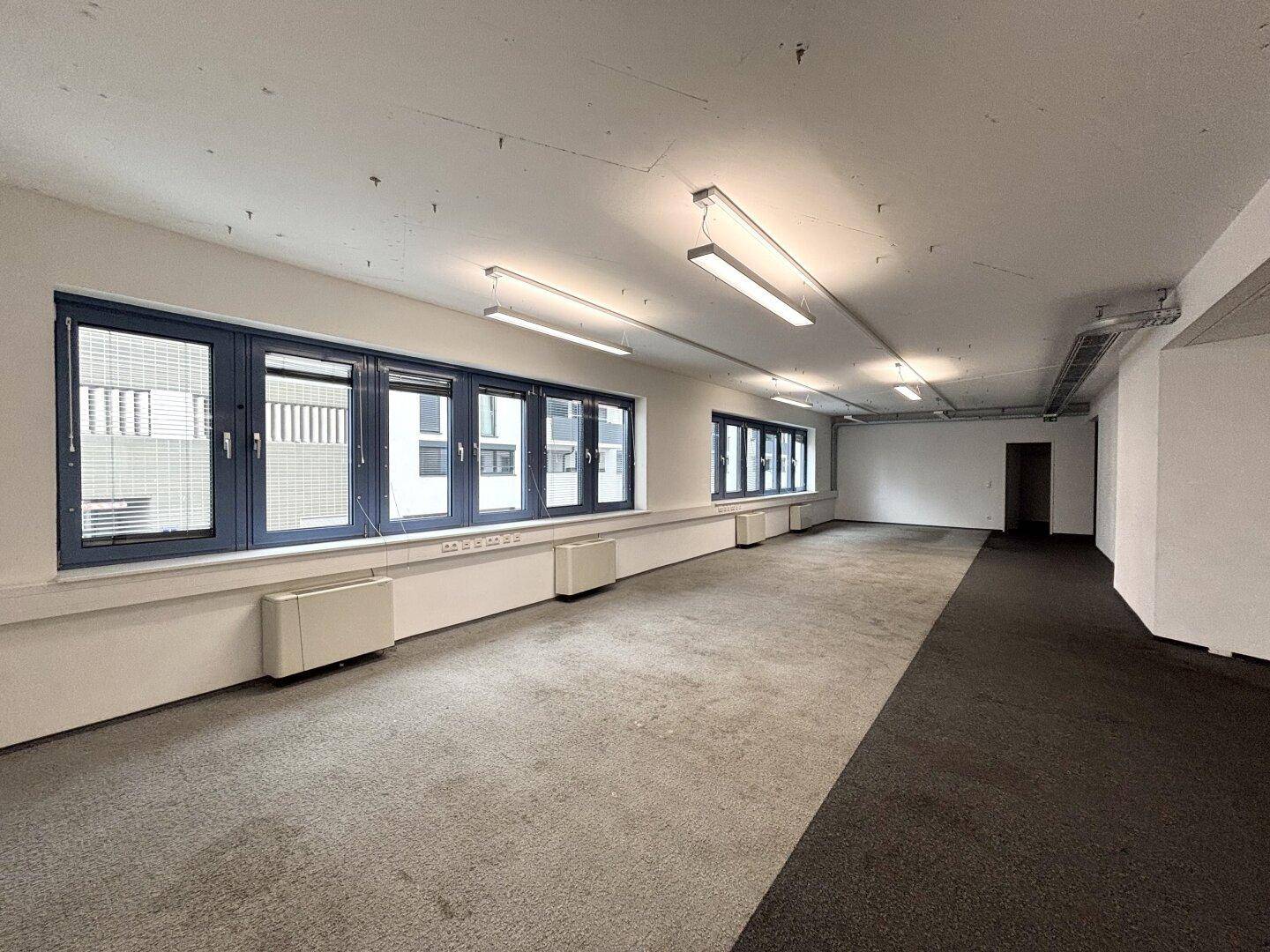 Heller Büroraum mit Teppichboden und Fenstern, die den Blick auf die Umgebung freigeben.