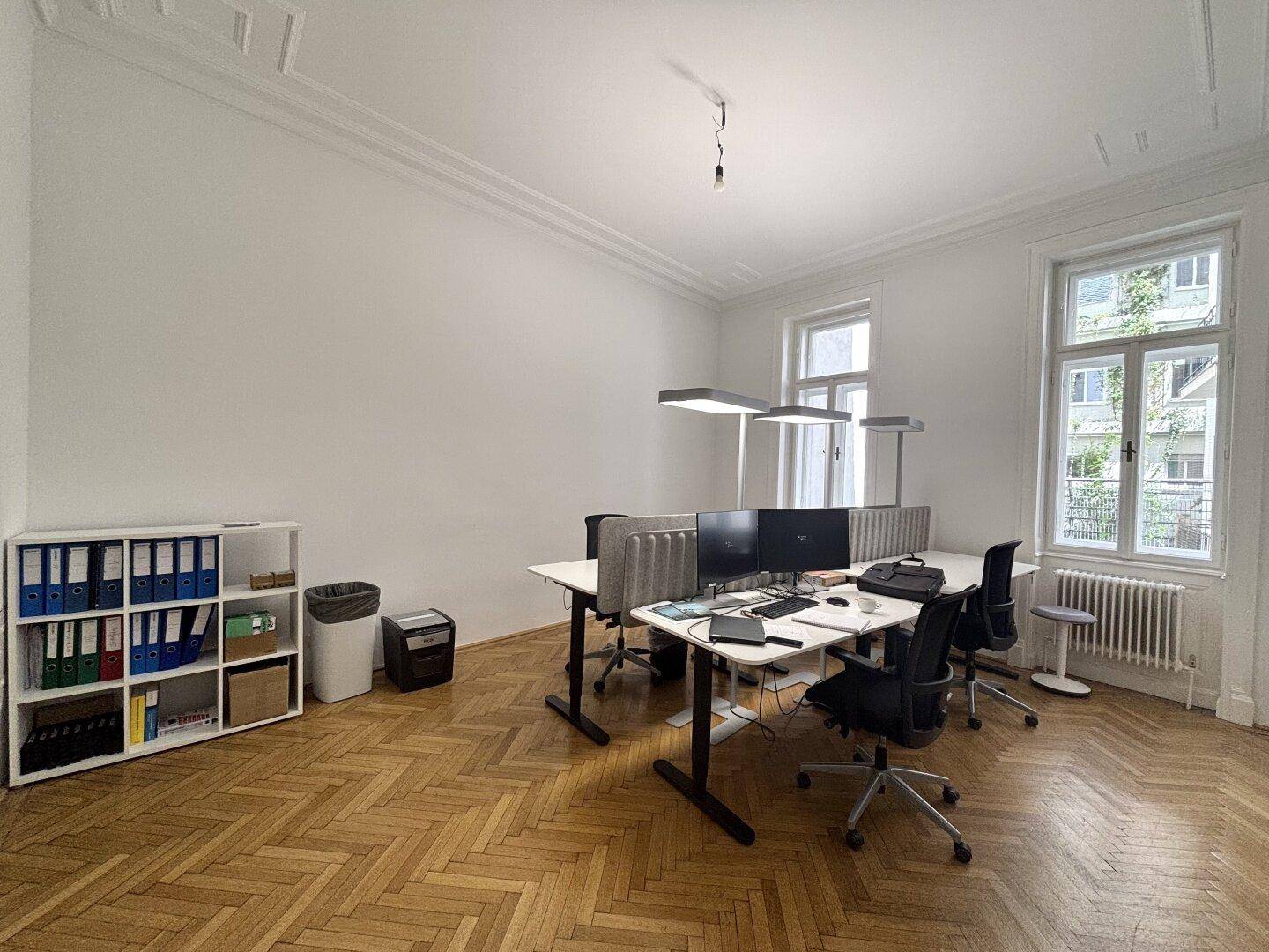 Geräumiges Büro mit Fischgrätparkett, mehreren Arbeitsplätzen, großen Fenstern und Regalen.