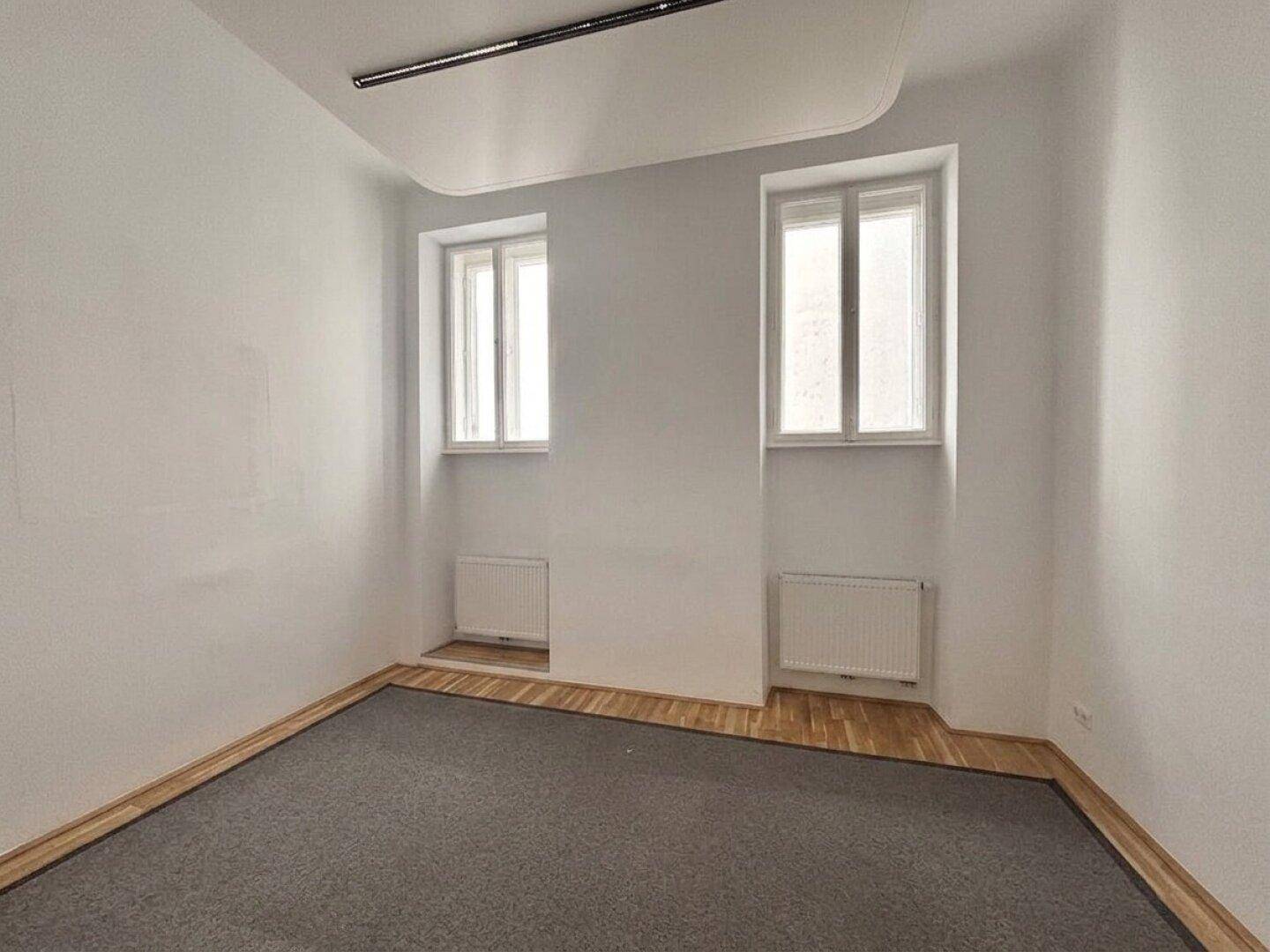 Leeres Büro mit Teppichboden, Holzboden am Rand und zwei hohen Fenstern.
